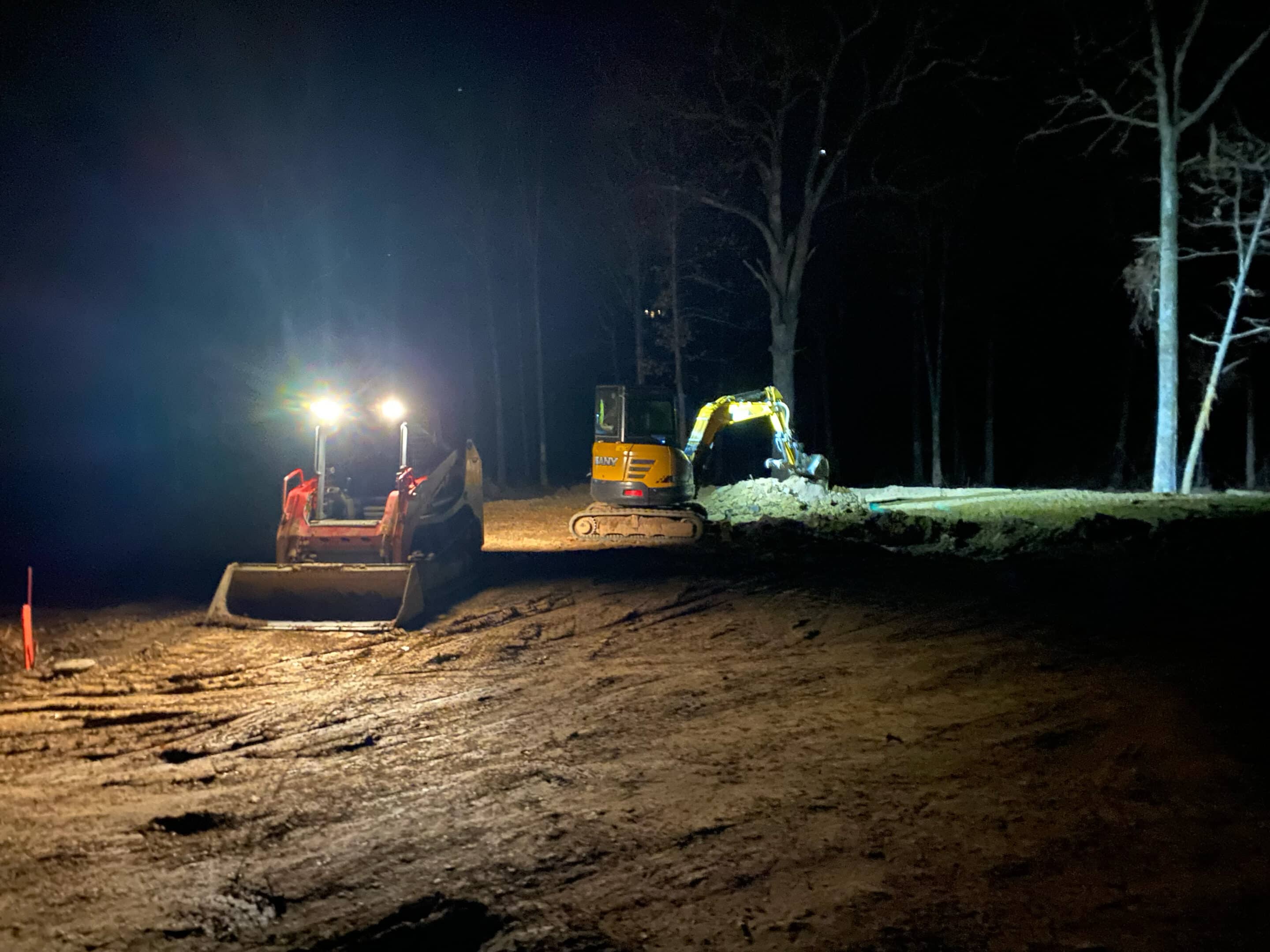 Two excavators operating at night in a dark outdoor area with bright work lights.
