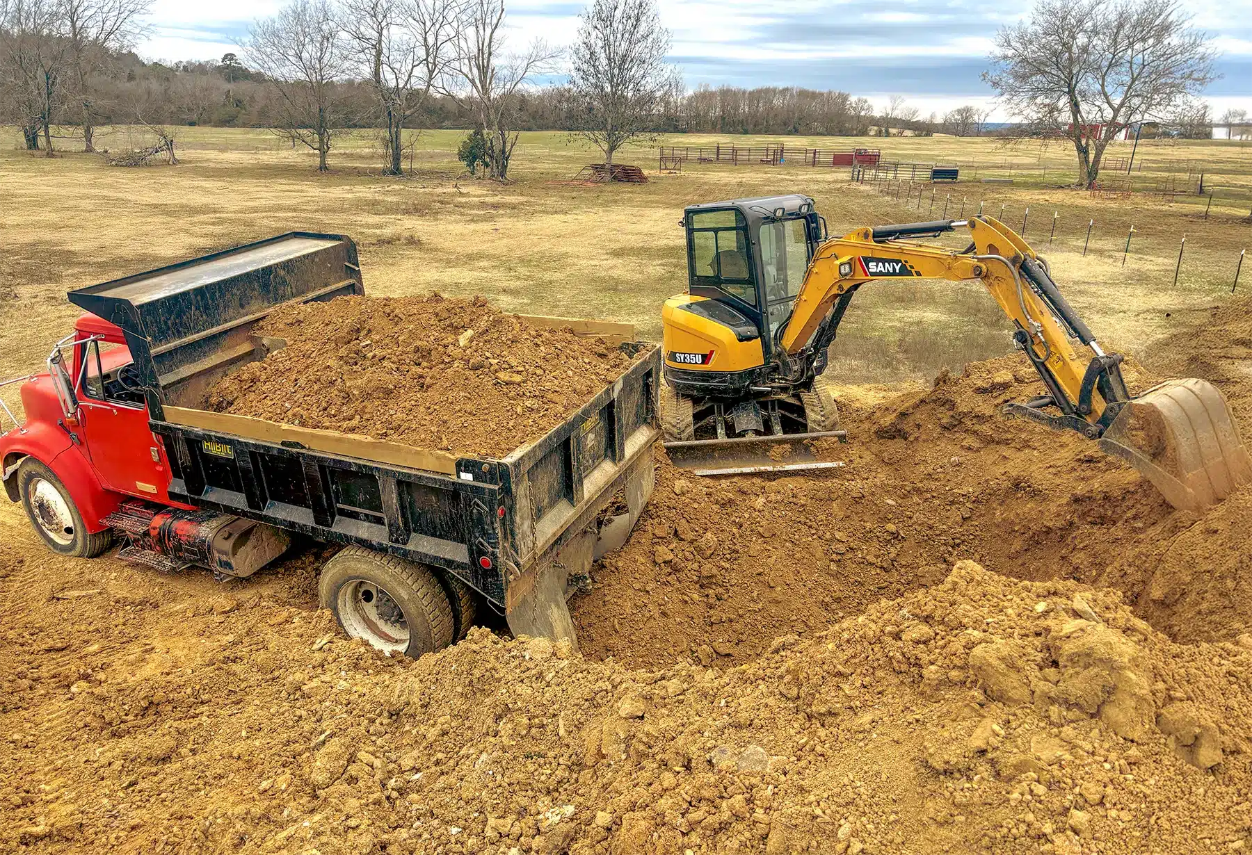 Excavator loading dirt into a truck on a construction site with a dirt mound.