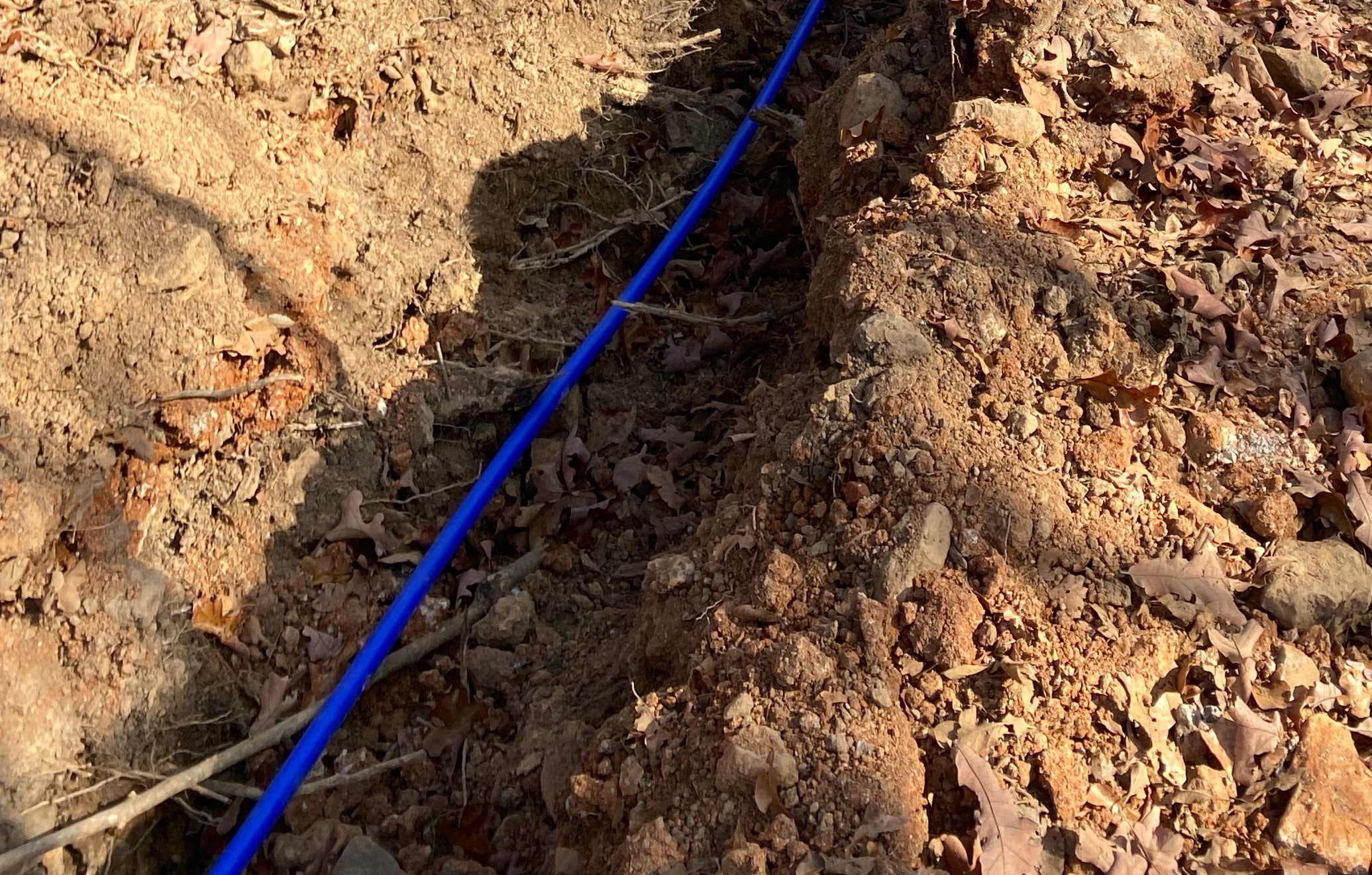 A blue drainage pipe laid in an excavated trench filled with loose soil and leaves.